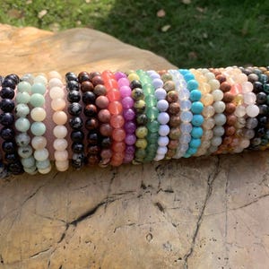 May include: A collection of beaded bracelets in various colors, including blue, black, pink, green, and orange. The bracelets are made of natural stones and are displayed on a light pink surface, with a natural stone background.