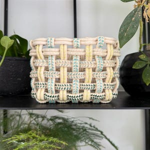 Può includere: Un cestino in ceramica quadrato caratterizzato da un design intrecciato realizzato con spessi cordoni di argilla arrotondati. La struttura alterna cordoni color crema tinta unita a cordoni decorati con piccoli motivi a foglia verde ottanio e macchie gialle.