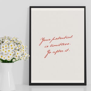 Könnte beinhalten: Gerahmter Druck mit dem Text "Your potential is limitless. Go after it." in roter Schrift auf cremefarbenem Hintergrund. Der schwarze Rahmen steht auf einer weißen Oberfläche neben einer weißen Vase mit Gänseblümchen.