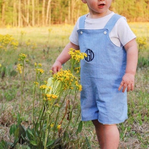 May include: A young child wearing light blue overalls with a white t-shirt underneath, standing in a field of yellow wildflowers. The overalls have a black monogram on the front. The background features a blurred view of trees.