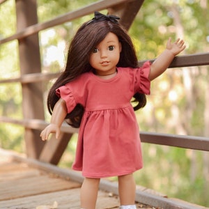 May include: A doll with long brown hair wearing a red dress and white sneakers. The doll is standing on a wooden bridge.