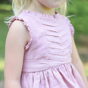 Puede incluir: Un vestido rosa pálido sin mangas con un escote con volantes y un corpiño plisado. El vestido está hecho de una tela de lino suave.