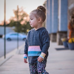May include: A young girl wearing a grey hoodie with a rainbow stripe on the pocket and black and white patterned trousers stands on a skateboard. She is wearing black and white trainers.