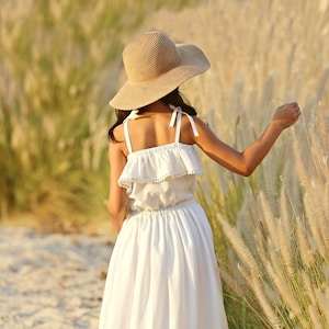 May include: A young girl wearing a white dress with a ruffled bodice and lace trim, standing in a field of tall grass. She is wearing a wide-brimmed straw hat.