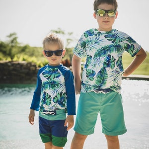 Op de afbeelding: Twee jongens dragen bijpassende zwemhemden met tropische print en zwembroeken. De jongen links draagt een shirt met lange mouwen en blauwe en groene zwembroeken. De jongen rechts draagt een shirt met korte mouwen en turquoise zwembroeken. Beide jongens dragen een zonnebril.