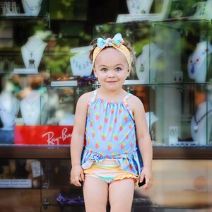 May include: A young girl wearing a blue and white swimsuit with a strawberry print and a yellow and blue headband. She is standing in front of a glass display case with jewelry.