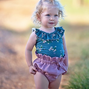 May include: A young child with blonde hair wearing a teal floral tank top and a pink ruffled skirt. The child is standing on a dirt path with green grass in the background.