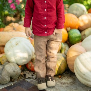 Op de afbeelding: Een jonge jongen in een rood overhemd met knopen en een bruine broek glimlacht terwijl hij voor een stapel pompoenen staat.