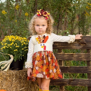 May include: A young girl wearing a white dress with a turkey design and a brown and yellow bow in her hair. She is standing in front of a wooden fence and holding onto it with her right hand. She is wearing yellow knee-high socks and brown boots.