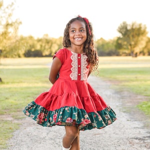 May include: A young girl wearing a red and green dress with a lace bodice and floral print skirt. She is smiling and walking on a dirt path.