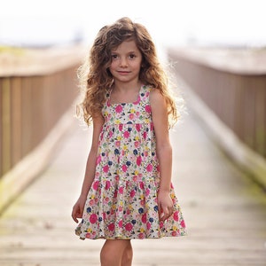 May include: A young girl with long curly hair is wearing a white sleeveless dress with a pink and yellow floral pattern. She is standing on a wooden walkway.