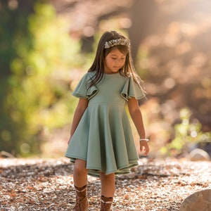 May include: A young girl wearing a sage green dress with ruffled sleeves and brown boots walks on a path covered in fallen leaves.