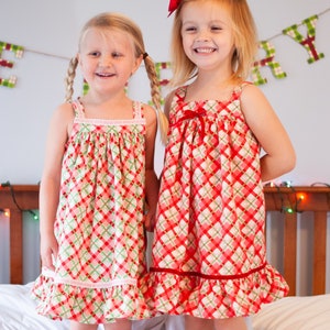 Pu&ograve; includere: Due ragazze che indossano abiti a quadri rossi e verdi con bordi bianchi e fiocchi rossi. Gli abiti hanno un orlo arricciato. Le ragazze sono in piedi su un letto bianco.