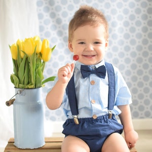 May include: A young child wearing a blue and white checkered shirt with suspenders and navy blue shorts. The child is holding a red lollipop and smiling. The child is sitting on a wooden crate.