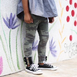 Könnte beinhalten: Ein Kind trägt ein hellblaues Jeansoberteil mit Rüschen am Saum und Camouflage-Leggings. Die Leggings haben einen Rüschen-Detail am unteren Ende und das Kind trägt schwarze Canvas-Schuhe mit weißen Schnürsenkeln.