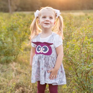 Könnte beinhalten: Ein junges Mädchen mit Zöpfen trägt ein weißes Shirt und ein grau-weißes Blumenkleid mit einer Eulen-Applikation. Sie trägt burgunderfarbene Strumpfhosen und braune Stiefel. Das Mädchen steht auf einem Feld mit hohem Gras.