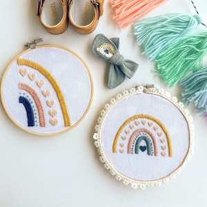 Op de afbeelding: Een verzameling handgemaakte babyartikelen. Inclusief een paar bruine leren babyschoenen, twee regenboog borduurringen, een grijze strik met een regenboogontwerp en een kwastjesslinger in pastelkleuren. De borduurringen tonen regenboogontwerpen met hartaccenten.