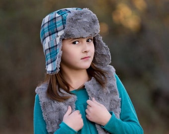 May include: A young person wearing a teal blue long-sleeved shirt with a gray faux fur vest. They are wearing a plaid and gray trapper hat with ear flaps.