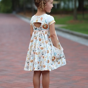 Puede incluir: Una niña pequeña lleva un vestido blanco con estampado de animales y globos de colores. El vestido tiene un escote en la espalda y un bolsillo grande en el lateral. Lleva sandalias blancas.