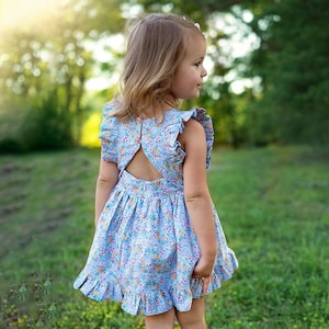 May include: A young girl wearing a light blue floral dress with a ruffled hem and a keyhole back. She is standing in a grassy field with green foliage.