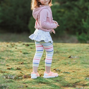 Può includere: Una bambina che indossa una felpa rosa con cappuccio, una gonna bianca con volant e leggings a righe. È in piedi in un campo erboso e guarda oltre la spalla.