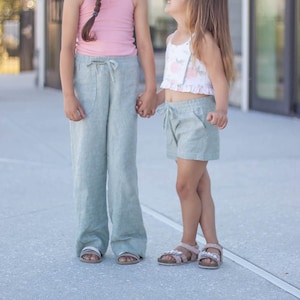 May include: Two girls wearing casual summer outfits stand outside. The girl on the left is wearing a pink tank top and light blue wide-leg pants. The girl on the right is wearing a white floral crop top and light blue shorts.