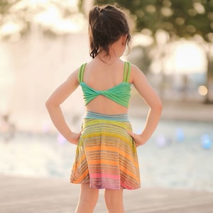 Op de afbeelding: Een jong persoon draagt een groen-regenboog gestreepte jurk met een twist detail op de rug. De jurk is gemaakt van een zachte, rekbare stof en heeft een zwierige rok. De persoon staat op een houten dek met een fontein op de achtergrond.