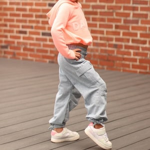May include: A young girl wearing a coral-colored hooded sweatshirt with the word "DA" and light blue cargo pants. She is wearing white sneakers with pink accents. The background is a brick wall and wooden deck.