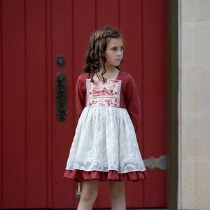 Puede incluir: Una niña pequeña con un vestido rojo y blanco con una capa de encaje y un patrón floral. El vestido tiene un escote cuadrado y mangas largas. La niña está de pie frente a una puerta roja.