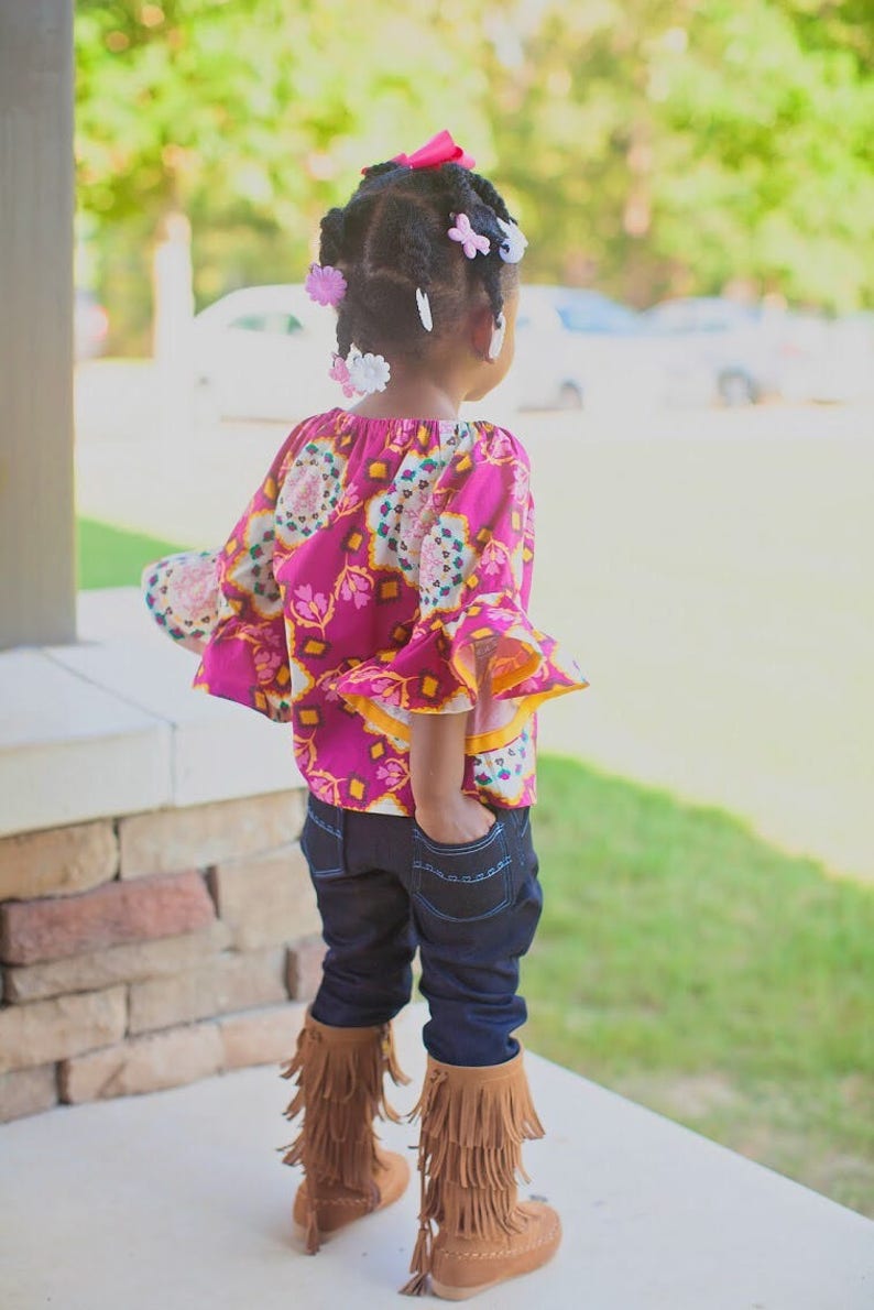 May include: A young girl wearing a pink and yellow patterned blouse with ruffled sleeves, dark blue jeans, and brown fringed boots. She has her hair styled in braids with pink and white hair clips.