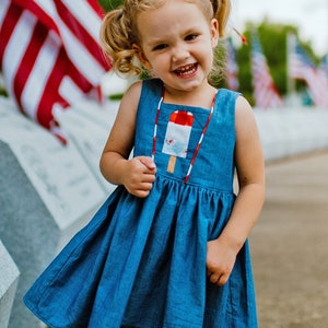 Puede incluir: Una niña pequeña con un vestido de mezclilla azul con un diseño de paleta de hielo roja, blanca y azul en la parte delantera. Ella está sonriendo y tiene el pelo en coletas. La niña está de pie frente a una bandera estadounidense.
