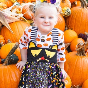 May include: A young girl wearing a black and white striped shirt with a black cat costume on top. The costume has a yellow and black cat face with a red nose and whiskers. The girl is standing in front of a pile of orange pumpkins.