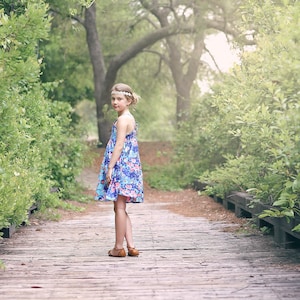 May include: A young girl wearing a floral print dress and a headband stands on a wooden walkway in a forest setting.