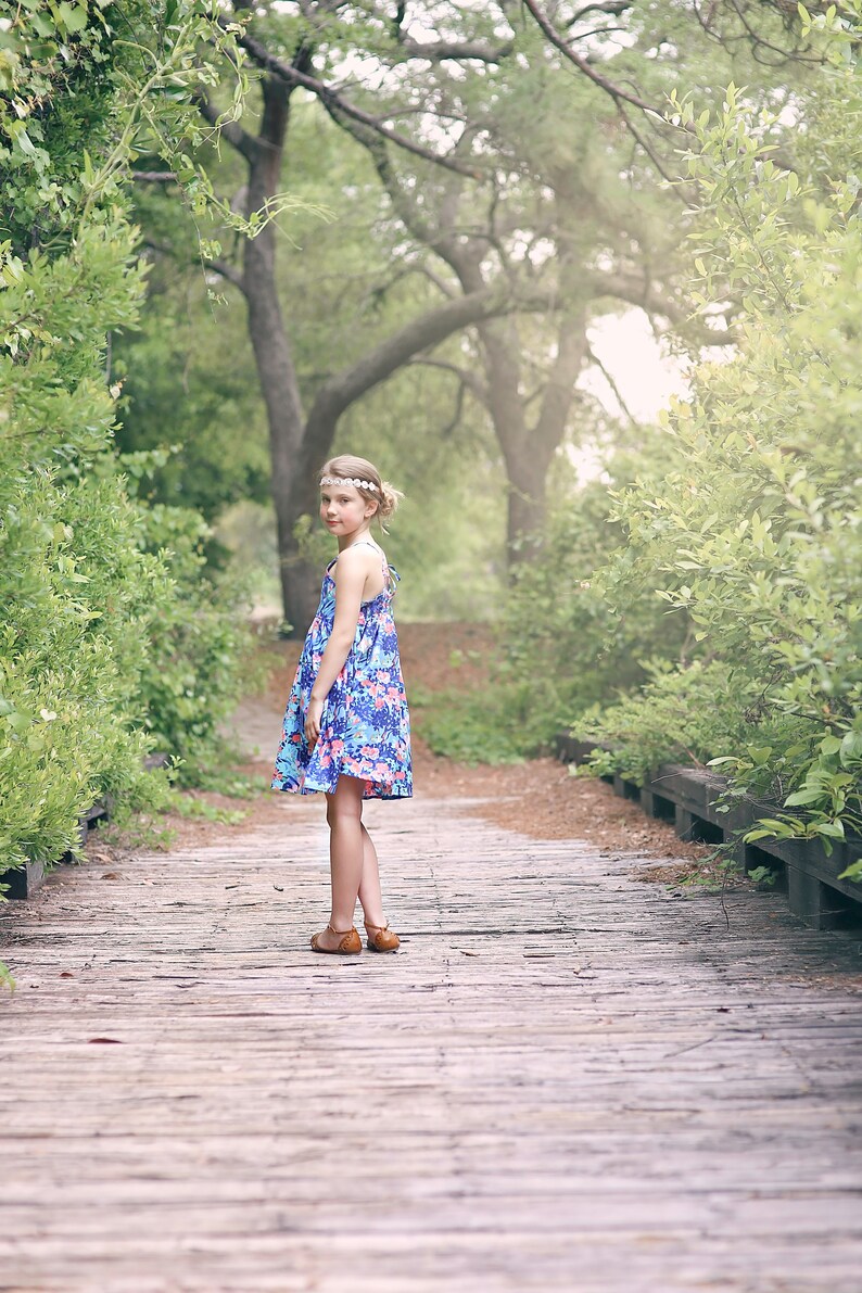 May include: A young girl wearing a floral print dress and a headband stands on a wooden walkway in a forest setting.