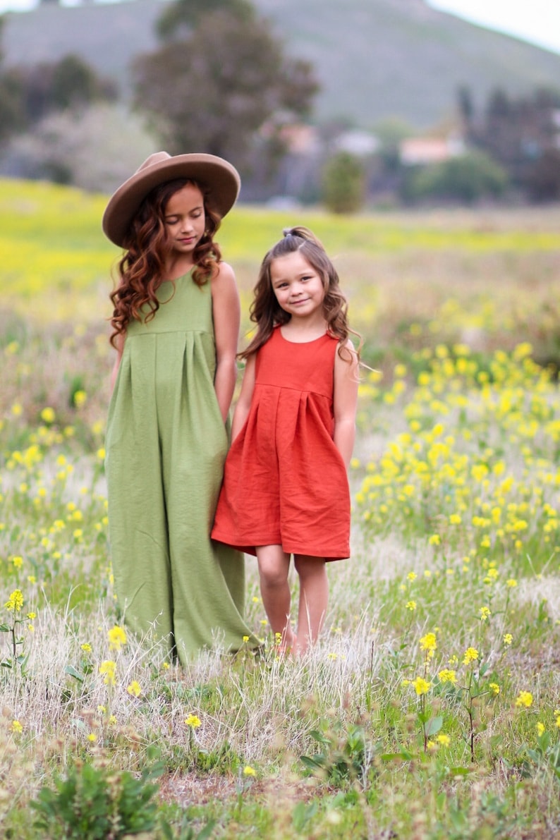May include: Two young girls wearing sleeveless dresses stand in a field of yellow wildflowers. One girl is wearing a green dress and a brown hat, while the other girl is wearing a red dress.