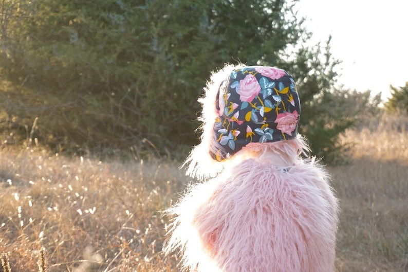May include: A young person wearing a pink fuzzy jacket and a floral patterned hat. The hat has a dark blue background with pink, yellow, and green flowers.