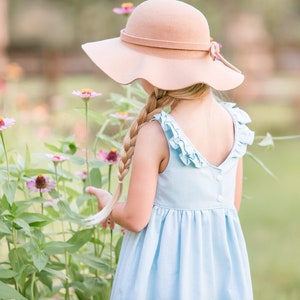 Puede incluir: Una niña pequeña con un vestido azul claro con volantes y un sombrero marrón de ala ancha, de pie en un campo de flores rosas.