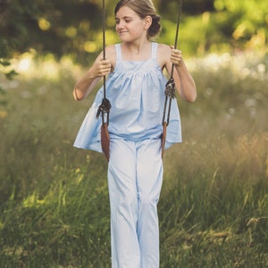 Pu&ograve; includere: Una giovane persona indossa una blusa azzurra larga senza maniche e pantaloni abbinati, seduta su un'altalena in un campo erboso.