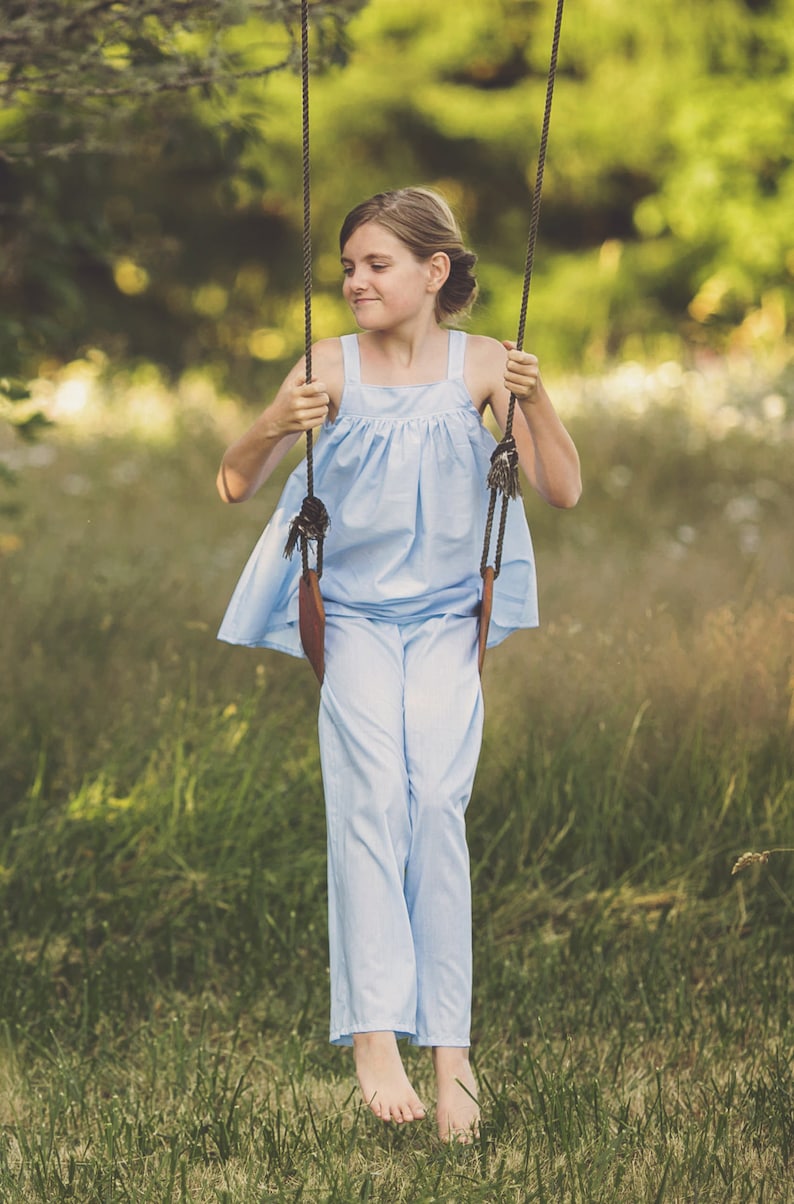 Pu&ograve; includere: Una giovane persona indossa una blusa azzurra larga senza maniche e pantaloni abbinati, seduta su un'altalena in un campo erboso.