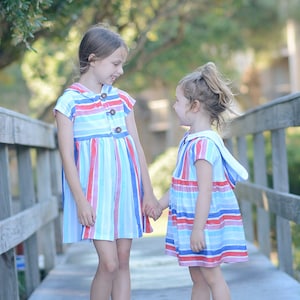May include: Two young girls wearing matching blue and red striped dresses with white accents stand on a wooden walkway. They are holding hands and looking at each other.