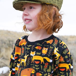 May include: A young person wearing a green, embroidered newsboy cap and a black sweater with a colorful woodland animal print. The sweater features foxes, owls, deer, bears, and hedgehogs.