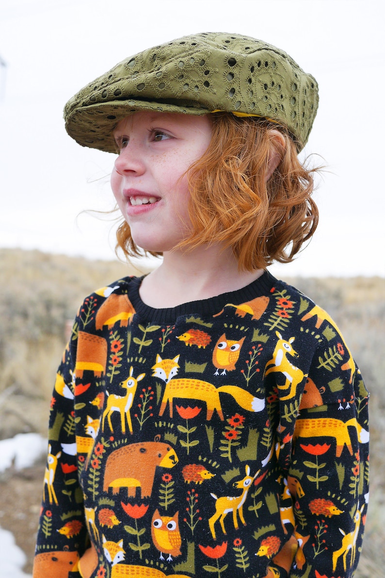 May include: A young person wearing a green, embroidered newsboy cap and a black sweater with a colorful woodland animal print. The sweater features foxes, owls, deer, bears, and hedgehogs.