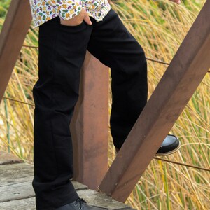 Op de afbeelding: Een persoon draagt een zwarte broek en een wit overhemd met knopen en een kleurrijk bladmotief. Ze staan op een houten brug met hun rechtervoet op de leuning. De persoon draagt zwarte leren laarzen.