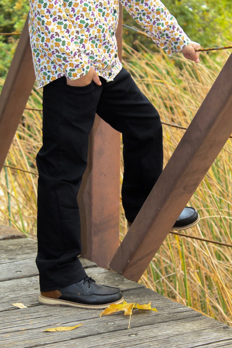 Op de afbeelding: Een persoon draagt een zwarte broek en een wit overhemd met knopen en een kleurrijk bladmotief. Ze staan op een houten brug met hun rechtervoet op de leuning. De persoon draagt zwarte leren laarzen.