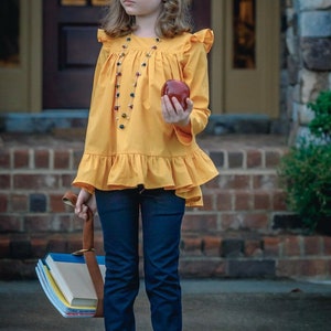 May include: A young girl wearing a yellow ruffled blouse with a beaded necklace, blue jeans, and sneakers. She is holding a red apple and a stack of books.