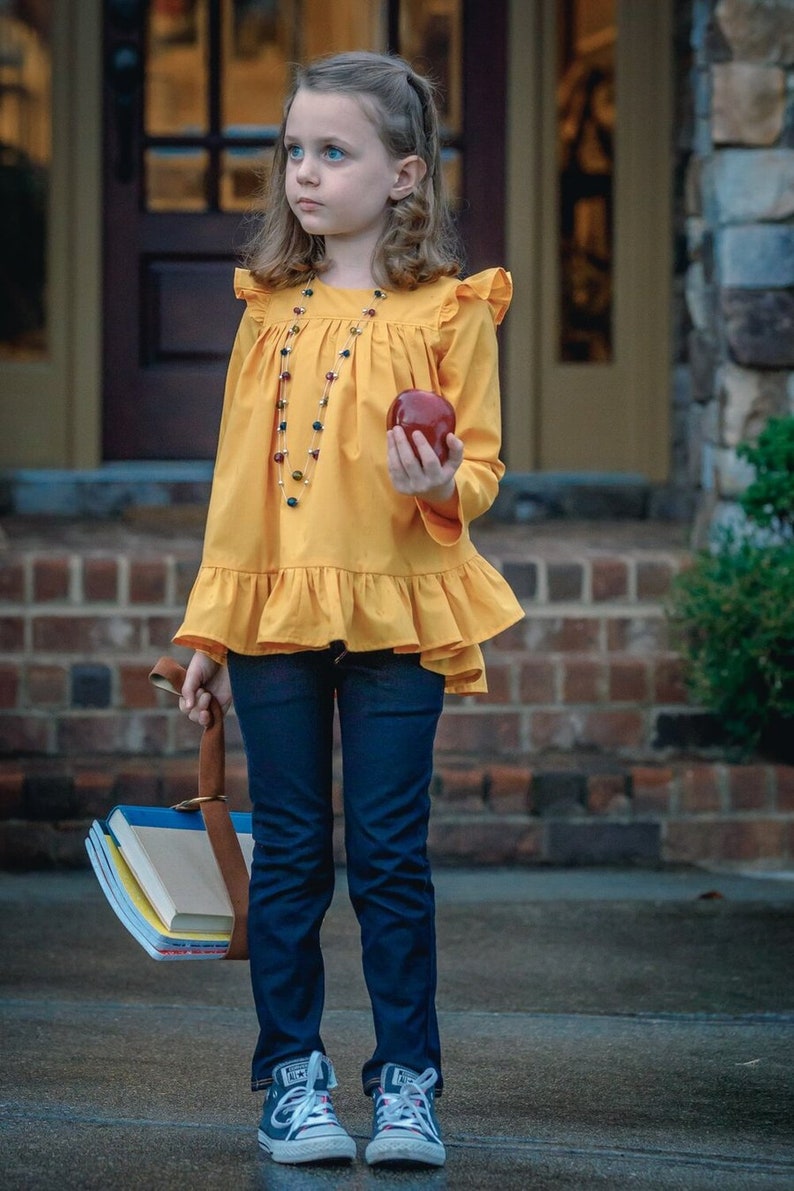 May include: A young girl wearing a yellow ruffled blouse with a beaded necklace, blue jeans, and sneakers. She is holding a red apple and a stack of books.