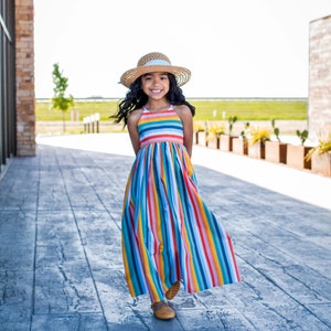 May include: A young girl wearing a rainbow striped maxi dress and a straw hat smiles while walking on a gray concrete patio.