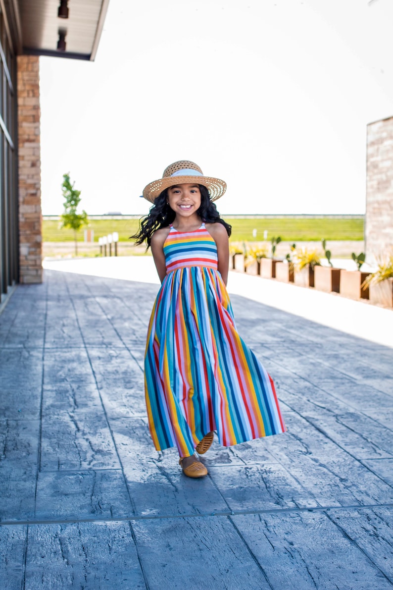 May include: A young girl wearing a rainbow striped maxi dress and a straw hat smiles while walking on a gray concrete patio.