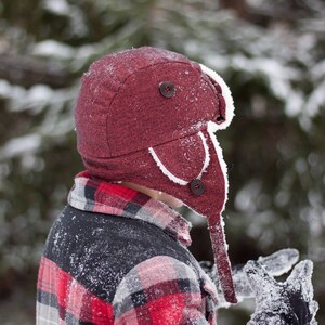 May include: A person wearing a red and black plaid flannel shirt with a red and white winter hat. The hat has a white fur trim and a button on the top. The person is standing in the snow.