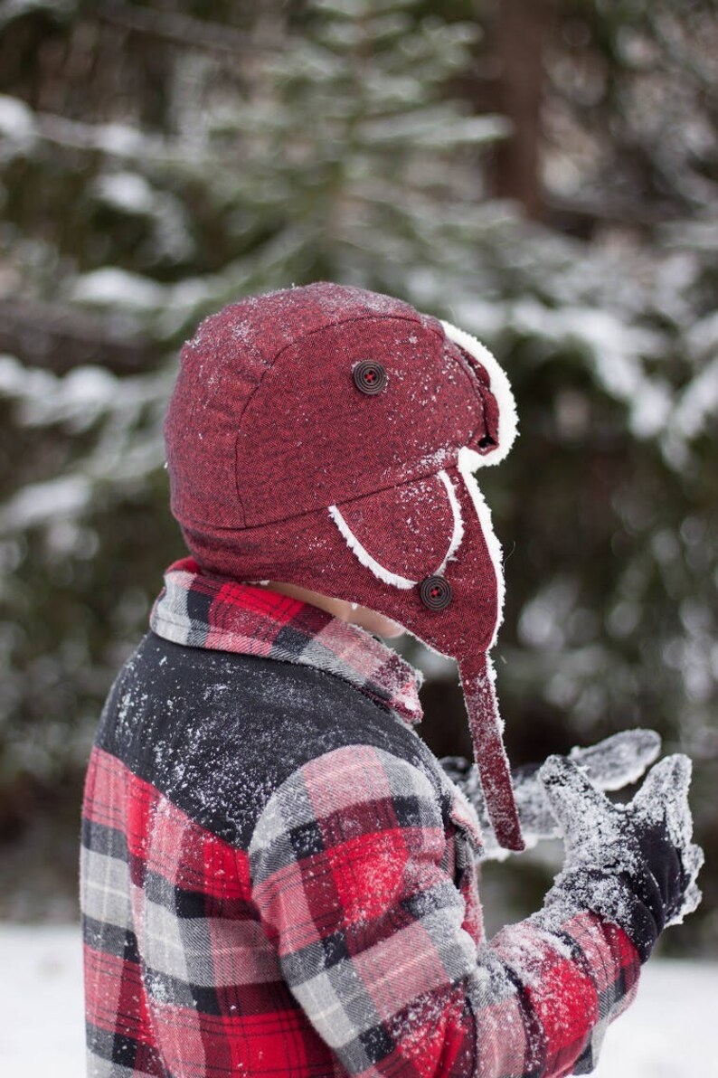 May include: A person wearing a red and black plaid flannel shirt with a red and white winter hat. The hat has a white fur trim and a button on the top. The person is standing in the snow.
